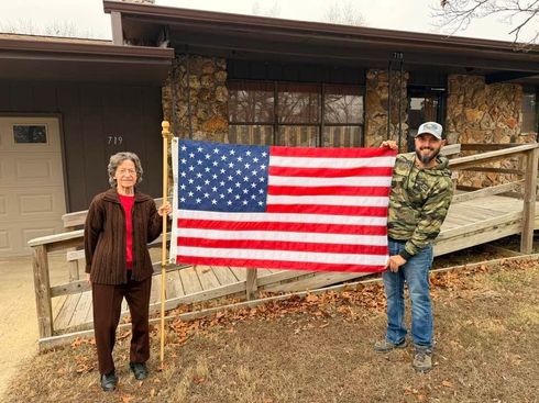 Two people standing outside holding a large American flag in front of a rustic house