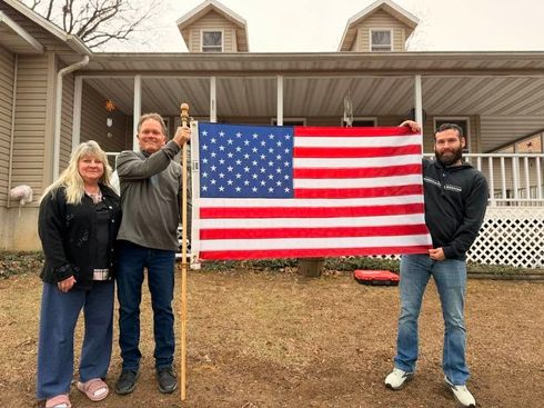 Three people hold a large American flag in front of a house porch.