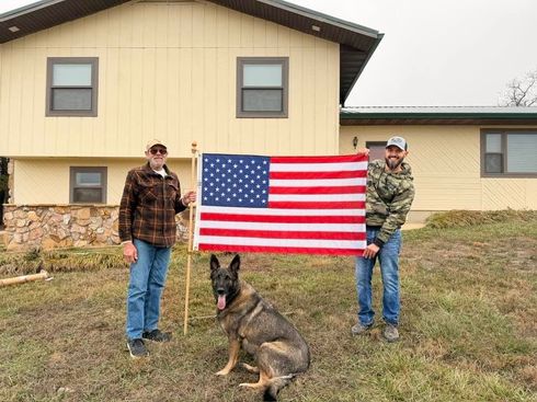 Two people holding an American flag with a German shepherd in front of a house