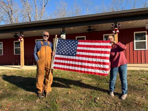 Two people hold an American flag outside a red building on a sunny day.