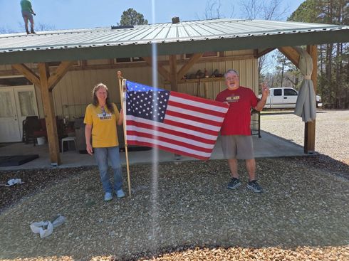 Two people standing under a shelter holding a large American flag in a gravel area