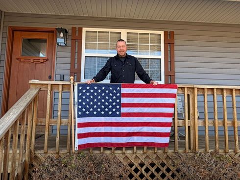 Man standing on a porch holding an American flag draped over the railing