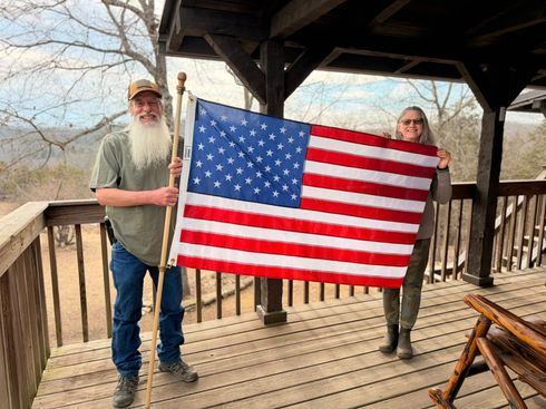 Two people holding a large American flag on a wooden porch with a rustic shelter and trees behind them