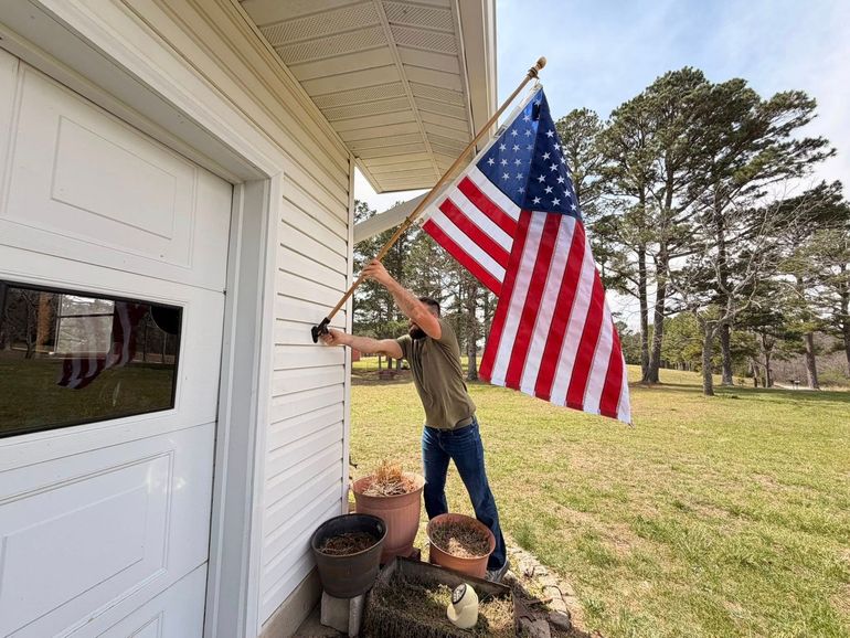 Person hanging a large American flag beside a white house on a sunny lawn.