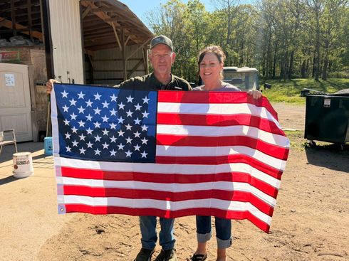 Two people holding a large American flag outdoors by a barn and trees