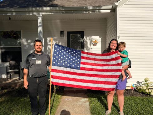 Three people holding a large American flag on a house porch in daylight.