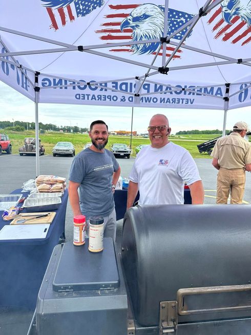 Two men smiling at a patriotic outdoor cookout booth with a grill, flags, and trays of food.