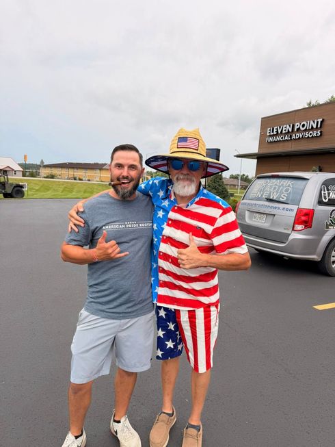 Two men posing outdoors; one wears an American flag outfit and cowboy hat, smiling beside a gray SUV.