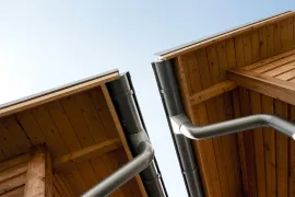 Looking up at two wooden roof eaves with silver gutters against a clear sky