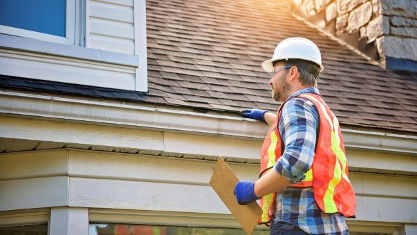 Worker in hard hat inspecting roof siding on a house with a clipboard.