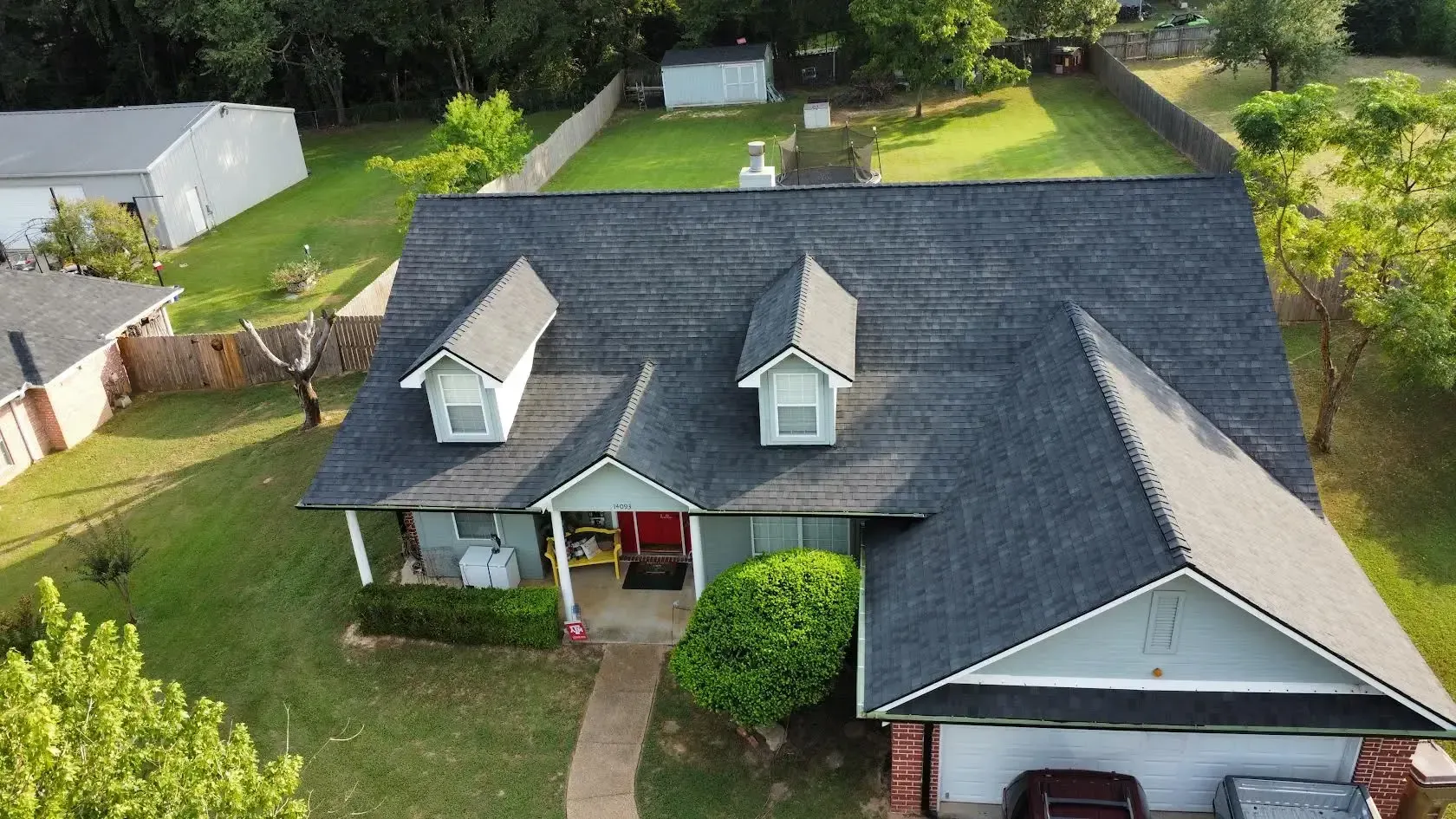 Aerial view of a gray-roofed suburban house with a driveway, front lawn, and surrounding trees.