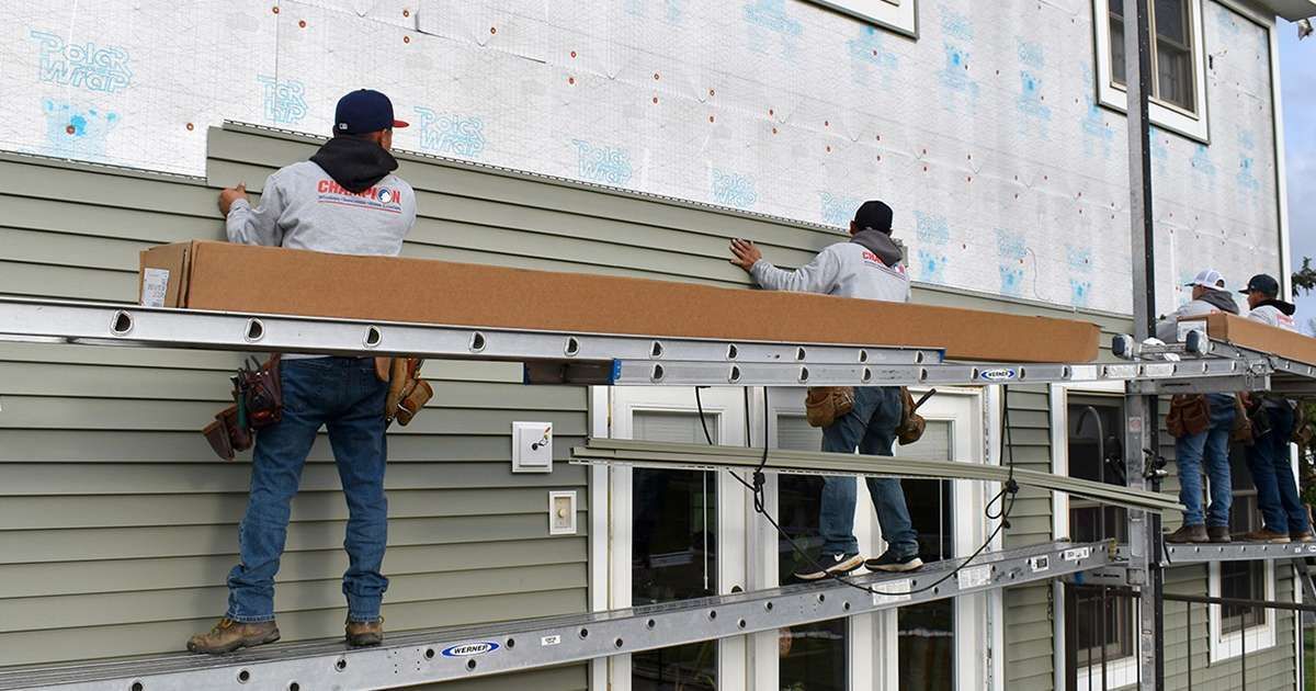 Workers installing long metal beams on scaffolding outside a building with a gray wall