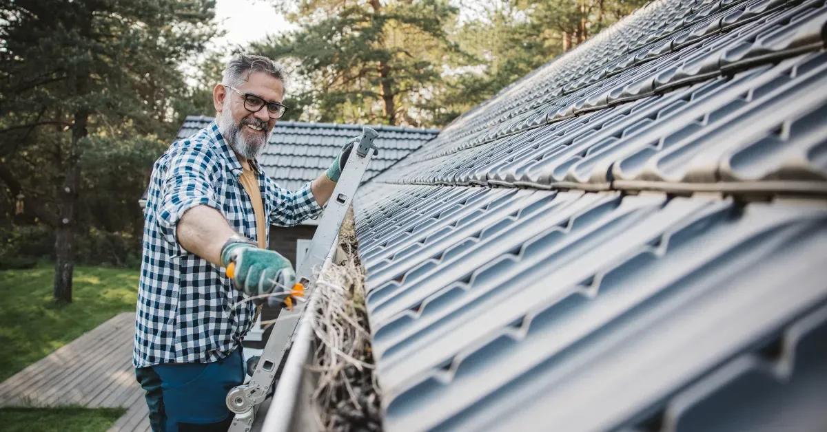 Man cleaning solar panels on a rooftop with a brush and hose, wearing gloves and glasses.