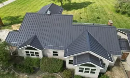 Aerial view of a house with a dark gray metal roof and large windows, surrounded by green lawn.