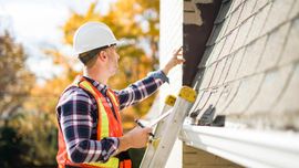Worker in hard hat and safety vest inspecting a house exterior on a ladder with tools.