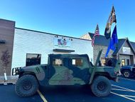 Camouflaged military Humvee parked outside a building with American flags and blue sky