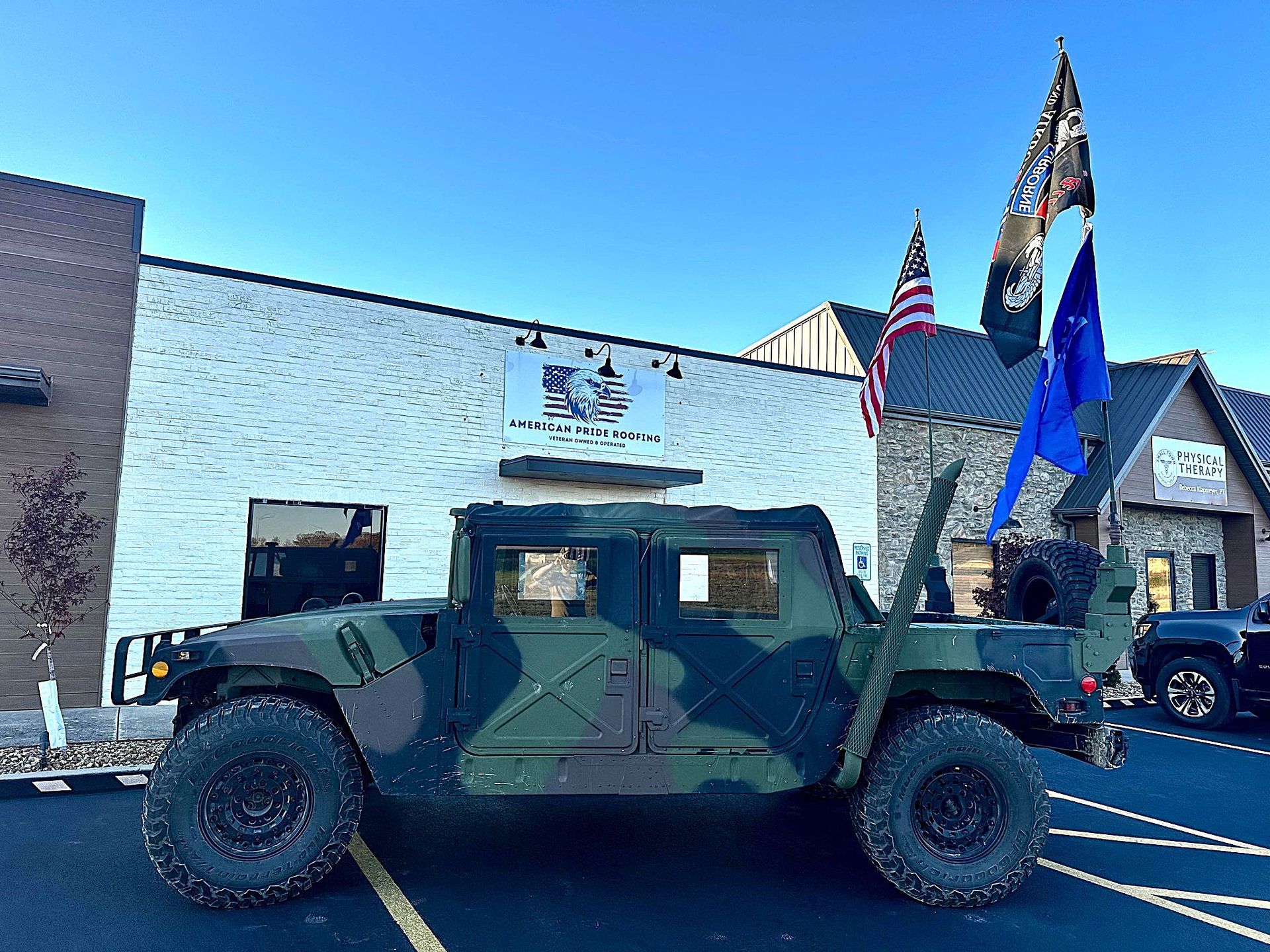 Camouflaged military Humvee parked outside a building with American flags and blue sky