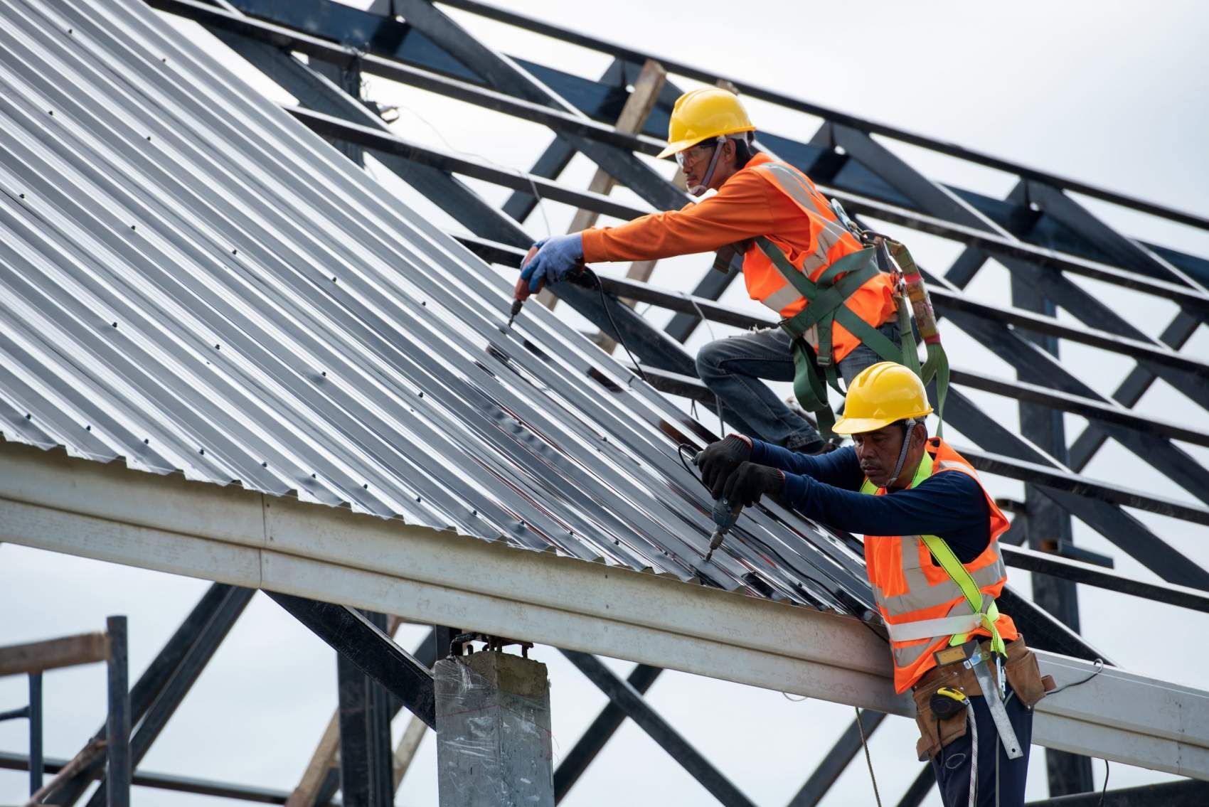 Two roofers in orange safety vests and helmets installing metal roofing on a building frame