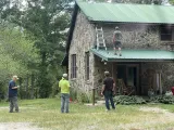 People on a ladder work on the green roof of a stone house in a wooded yard.