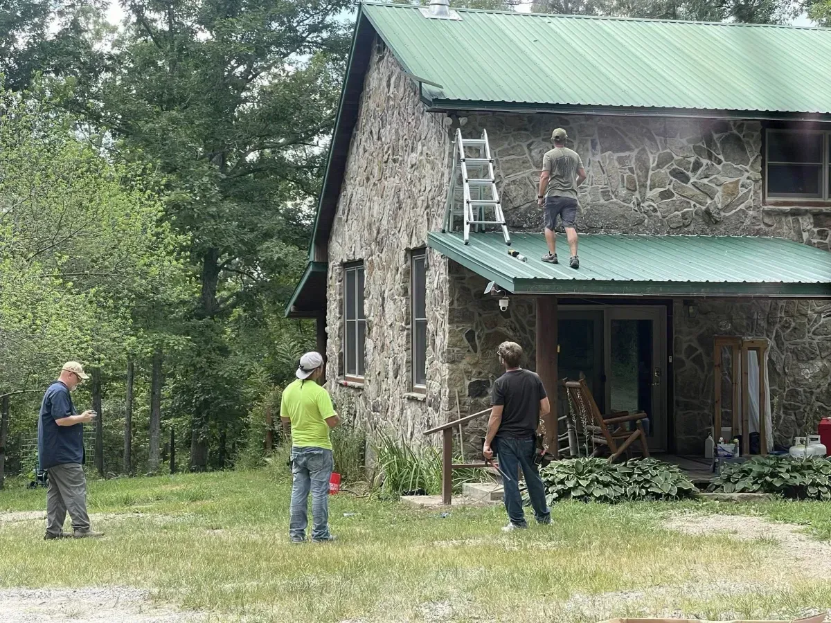 People on a ladder work on the green roof of a stone house in a wooded yard.