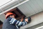 Worker in orange hard hat using a drill under a roof eave