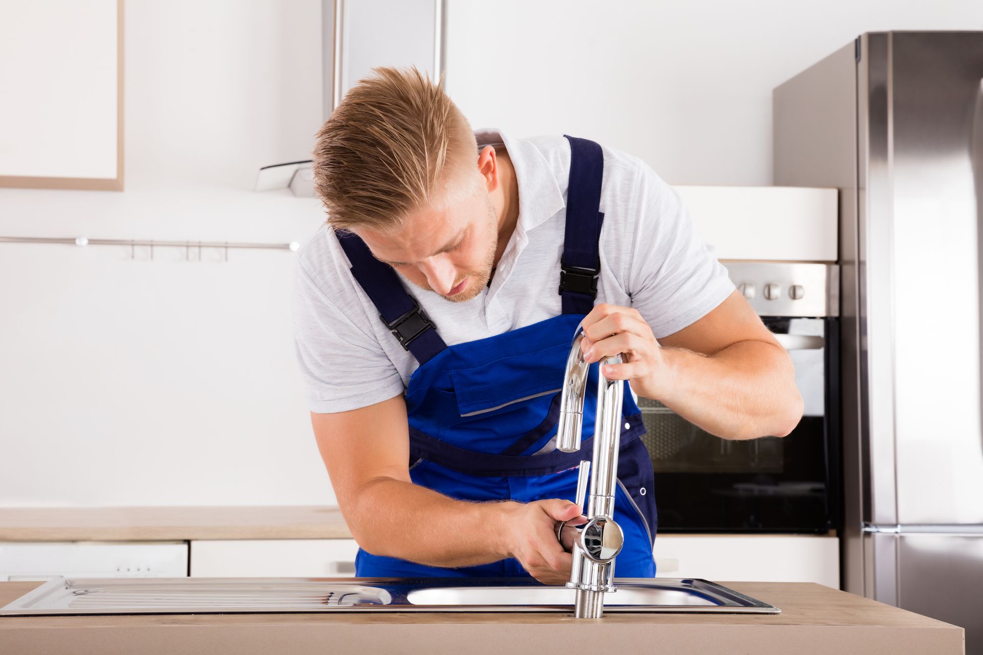 A plumber in blue overalls fixes a kitchen faucet, focusing intently. He's in a modern kitchen with white cabinets.