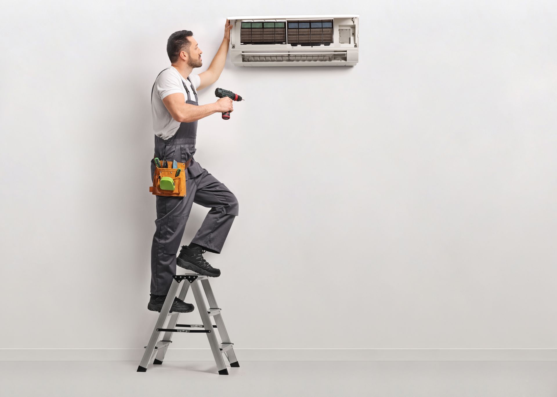 An HVAC technician on a stepladder using a drill to service a wall-mounted air conditioning unit. An HVAC technician on a stepladder using a drill to service a wall-mounted air conditioning unit.