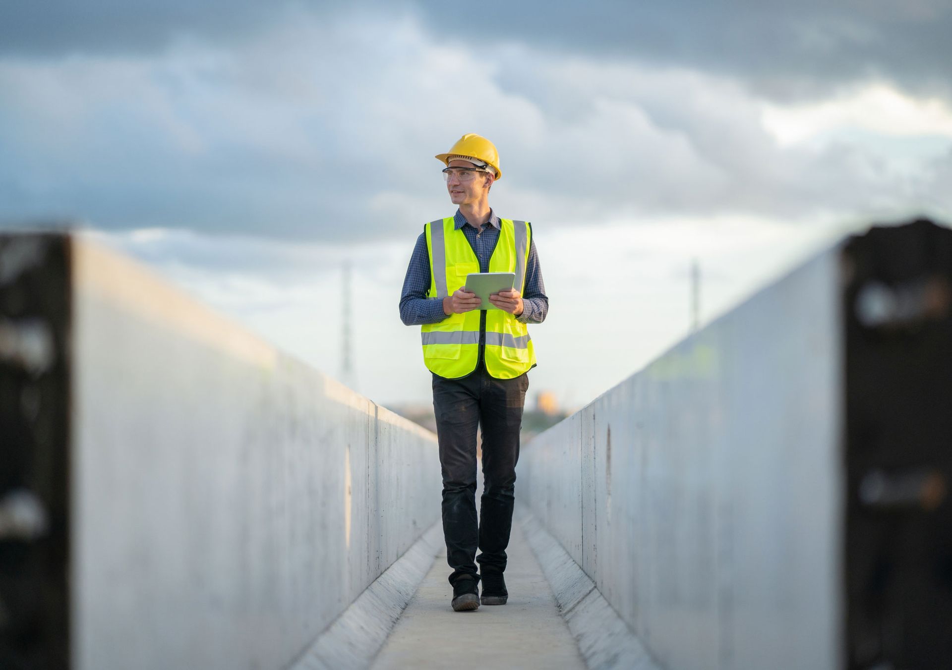 Construction worker in a yellow vest and helmet holding a tablet on a site. Construction worker wearing safety gear, walking and looking at a tablet on a concrete structure, overcast sky.