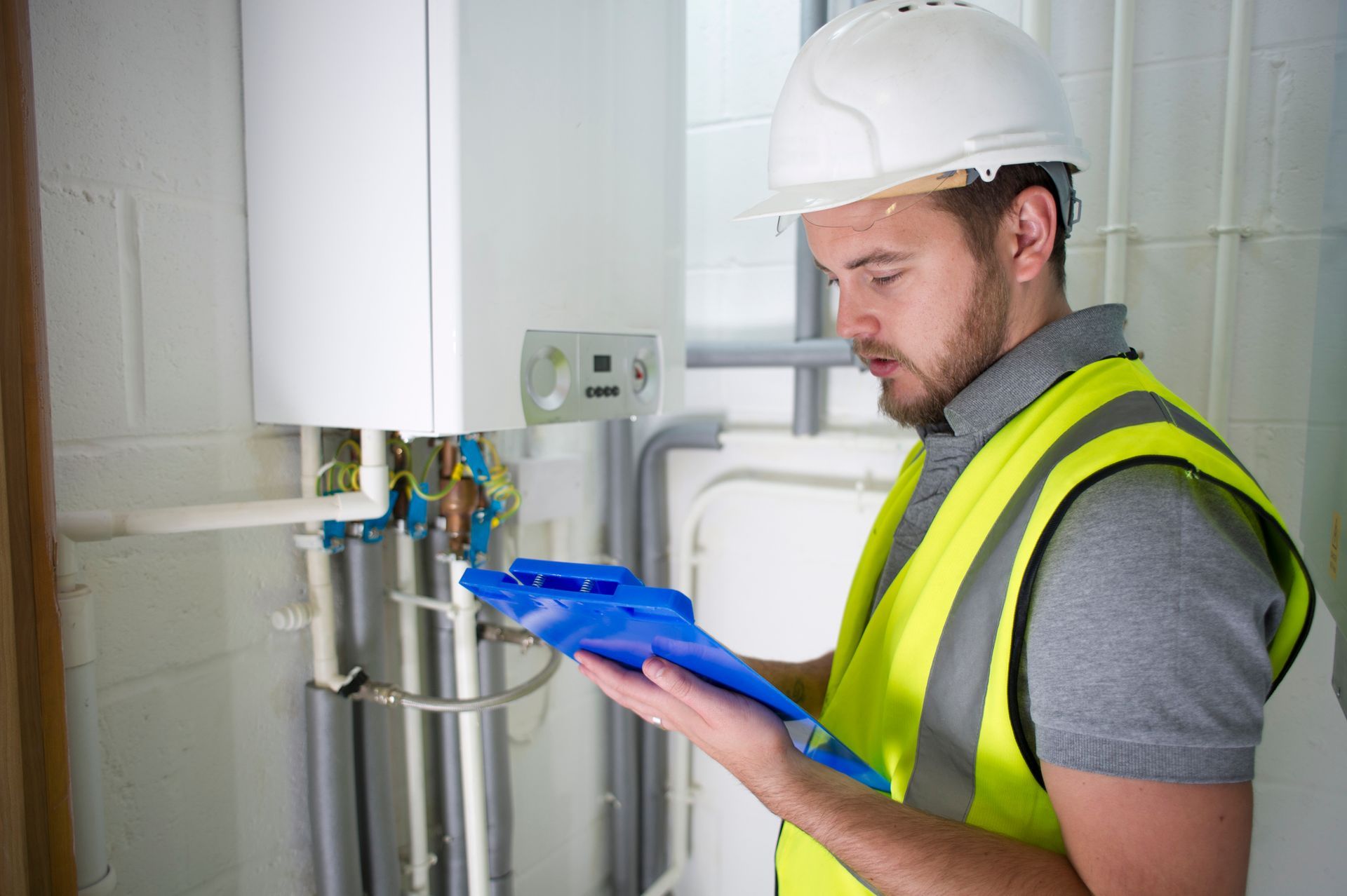A man in a safety vest and hard hat examines a boiler. He holds a blue clipboard, looking intently.