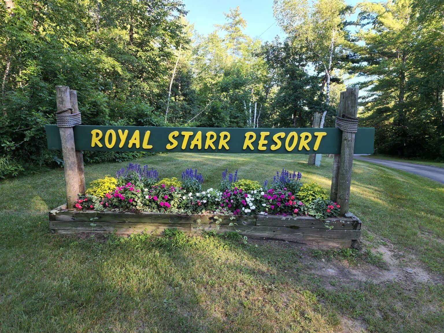 Wooden sign reading “ROYAL STARR RESORT” with flowers, beside a grassy roadside under trees