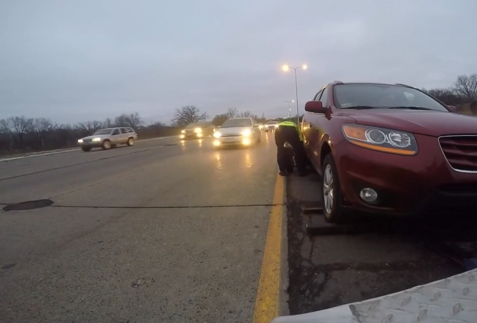 Person changing a tire on a red SUV parked on the side of a highway. Evening.