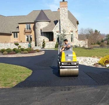 Person operating a yellow road roller on a newly paved driveway in front of a stone house