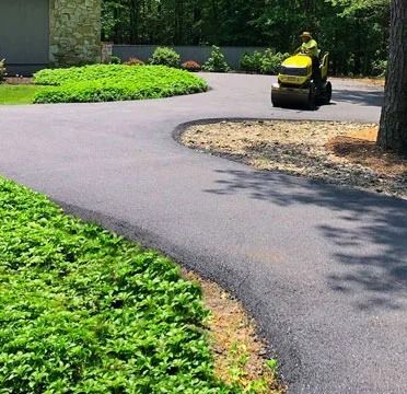 Yellow ride-on mower on a curving paved path beside green landscaping