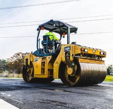 Yellow road roller compacting a wet asphalt road with an operator under a canopy