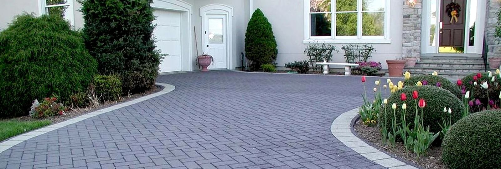 Landscaped front yard with a curved paver driveway leading to a white house and flowering garden beds