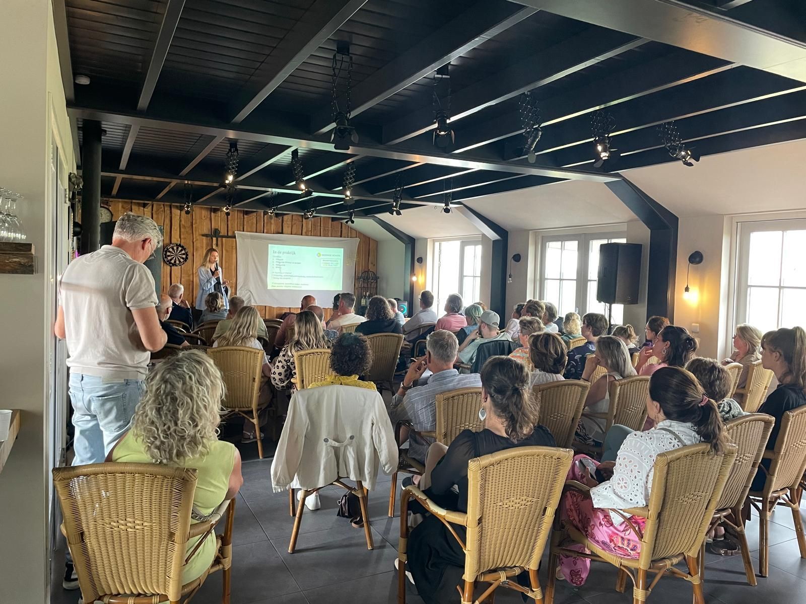 Mensen zitten bij een presentatie in een lichte ruimte; een spreker staat vooraan en een projectiescherm is zichtbaar.