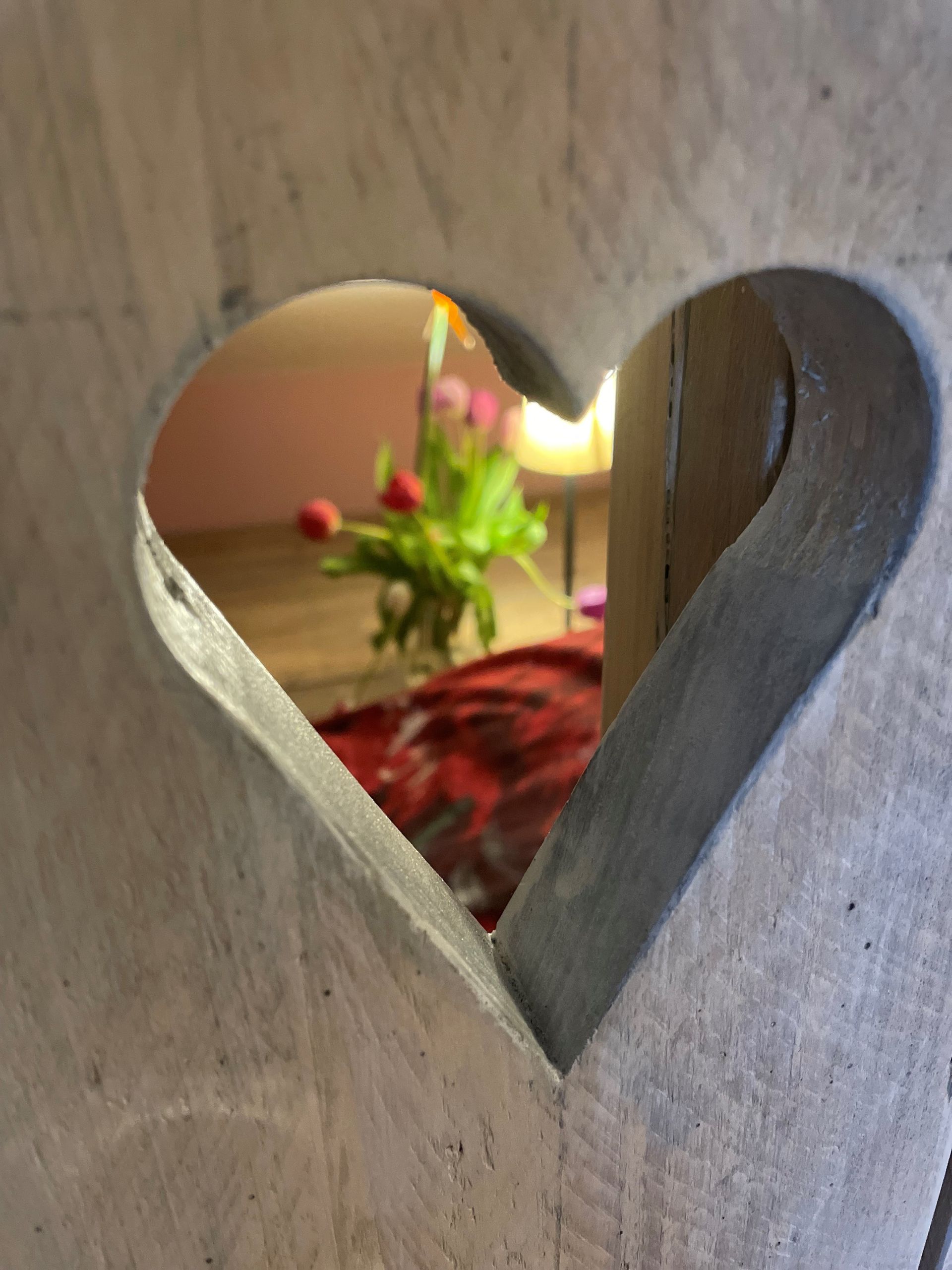 Een hartvormige uitsparing in een houten oppervlak omlijst een wazig beeld van een kamer vol bloemen.