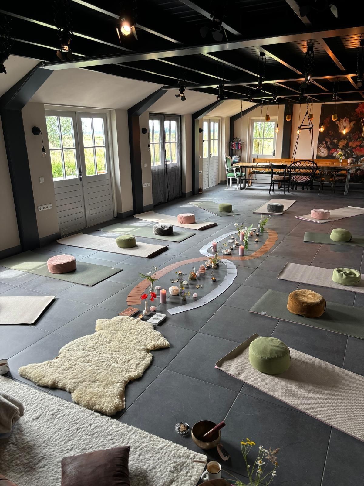 Yogastudio met matten, kussens en decoraties op een donkere vloer. Door de ramen komt natuurlijk licht binnen.