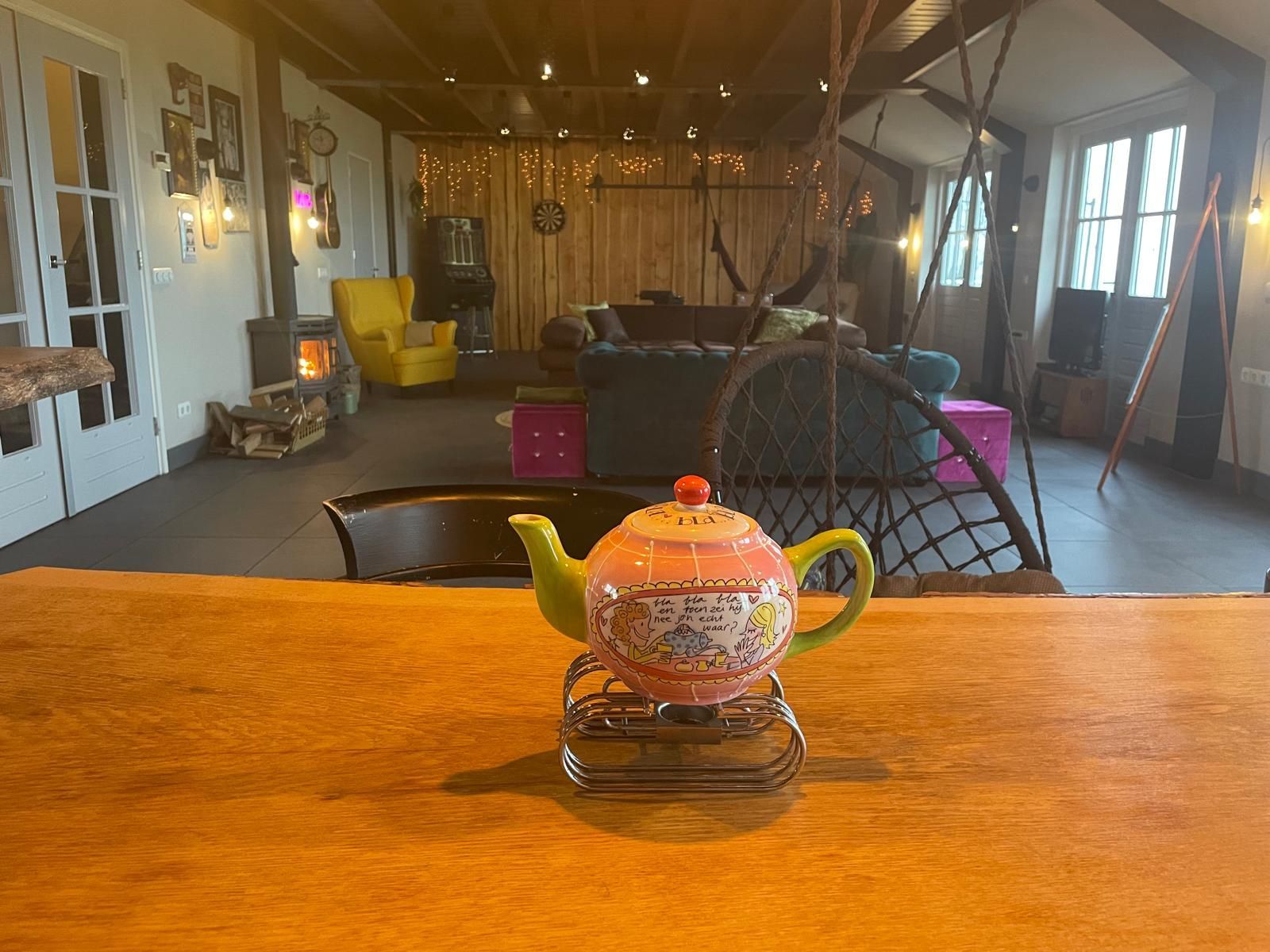 Een kleurrijke theepot op een houten tafel, met op de achtergrond een gezellig interieur met open haard, bank en sfeerverlichting.