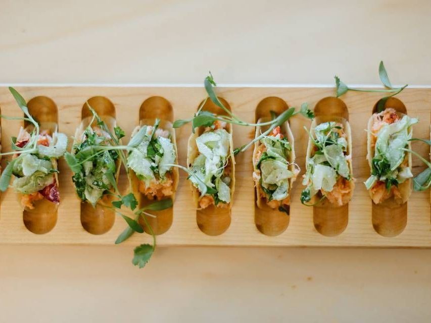 Taco tray with small tortillas topped with shredded lettuce, herbs, and sauce on a wooden board