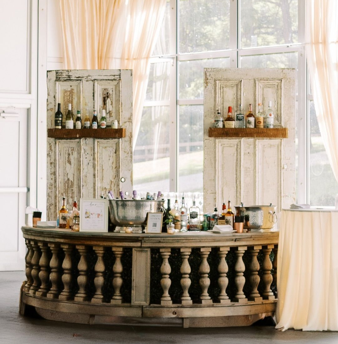 Rustic bar setup with bottles and glassware in a bright room by large windows