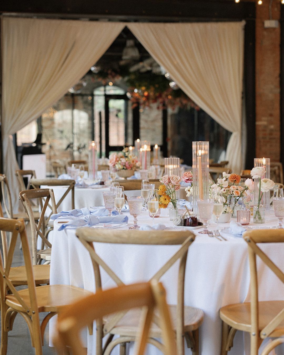 Elegant wedding reception tables with white linens, candles, and wooden chairs in a softly draped venue