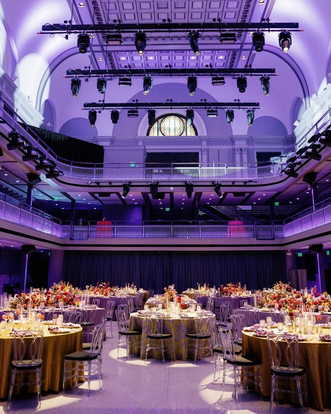 Purple-lit banquet hall with round tables set for a formal event beneath an ornate balcony and ceiling lights