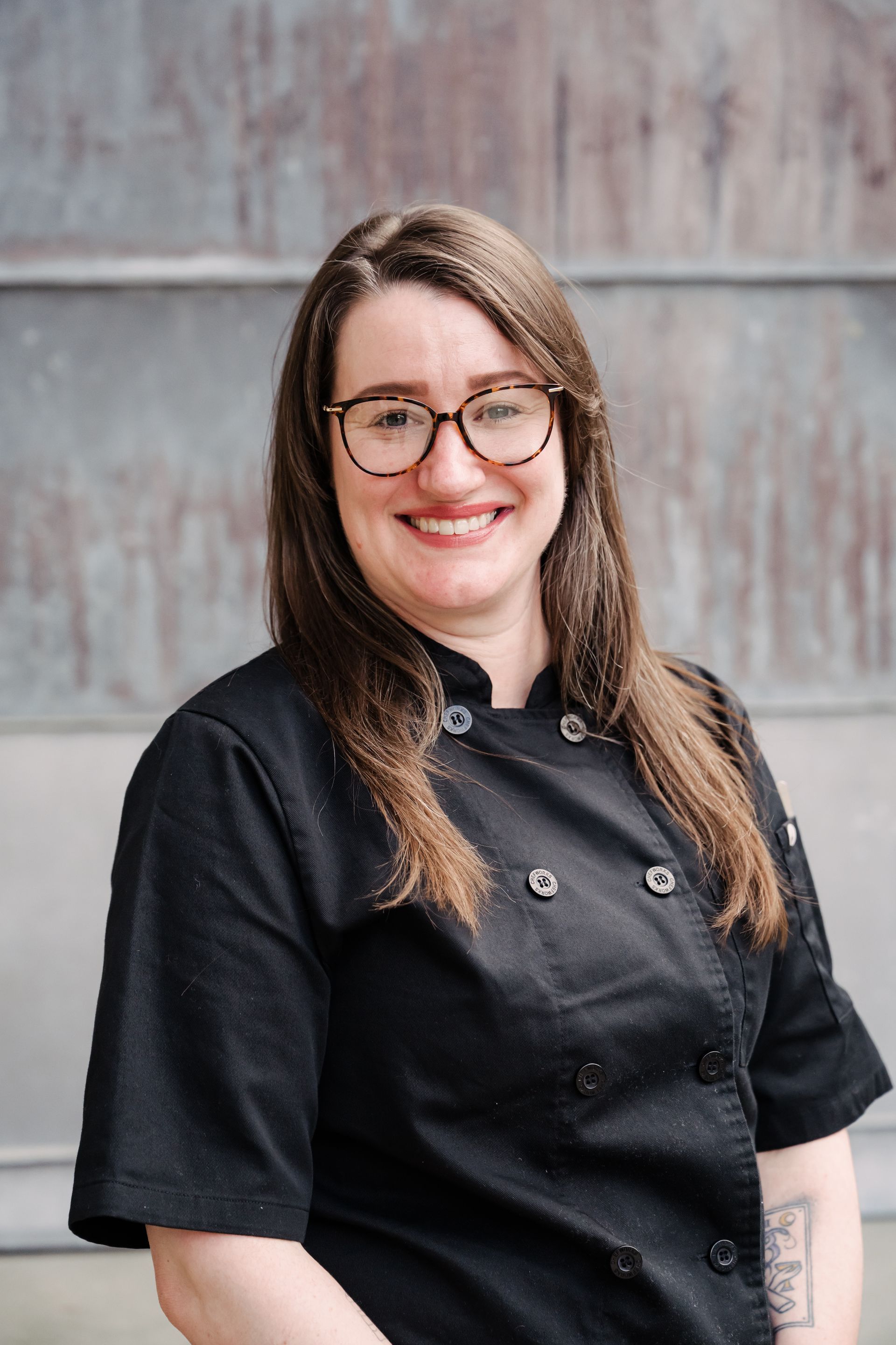 Smiling person in a black chef coat with glasses, standing against a gray wall.