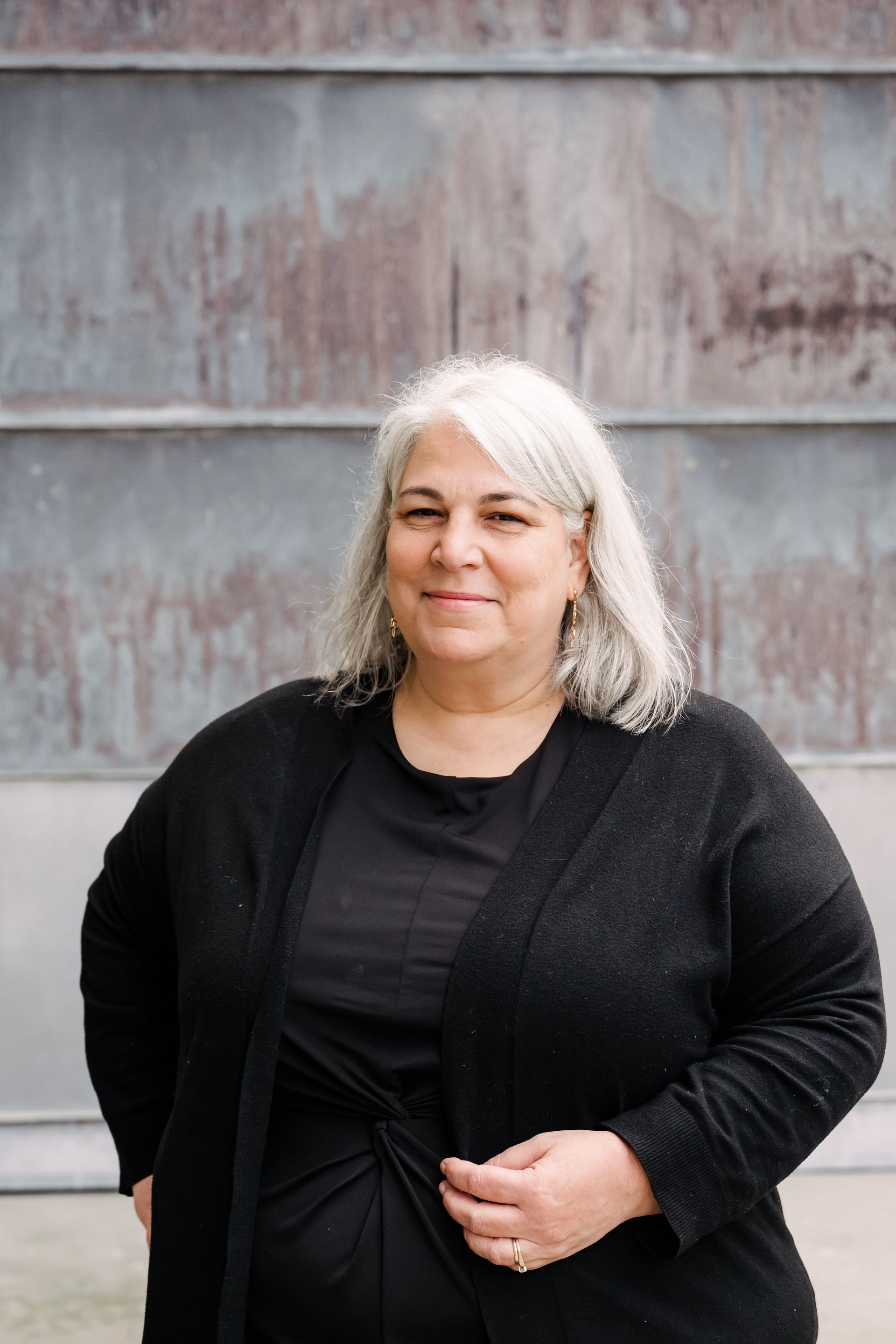 Woman in black jacket standing against a weathered wall, smiling slightly.