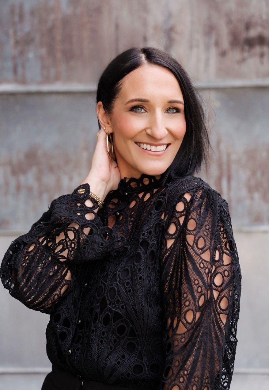 Smiling person in a black lace top, hand near neck, standing against a weathered wall.