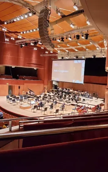 Empty concert hall with a stage, orchestra seating, and a large projection screen under warm wood ceiling lights