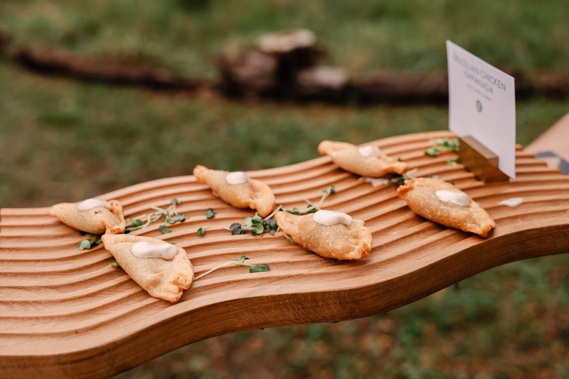 Wooden tasting board with small pastries and herbs, outdoors with a blurred green background