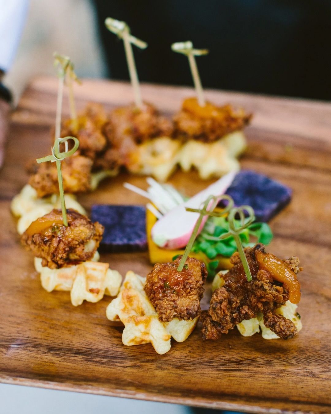 Assorted bite-sized skewers and fried appetizers on a wooden serving board, garnished with greens and flowers