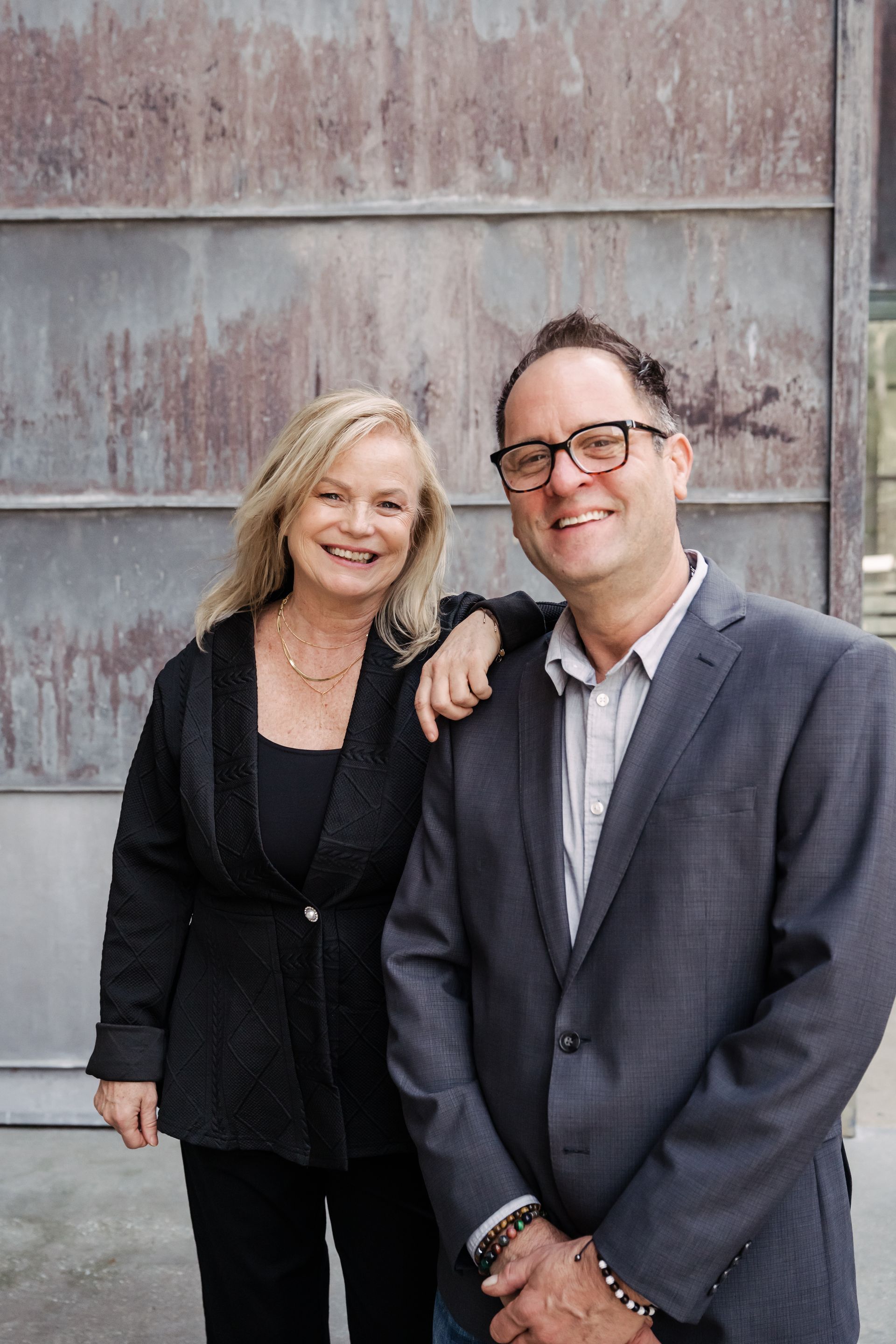 Two smiling people standing together against a weathered metal wall.